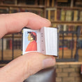 Load image into Gallery viewer, Hand holding a small book with a portrait, against a blurred bookshelf background