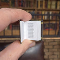 Load image into Gallery viewer, Hand holding a small open book with a blurred bookshelf background