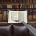 Load image into Gallery viewer, Open book held by tweezers against a blurred bookshelf background