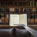 Load image into Gallery viewer, Open book held by tweezers in front of a blurred bookshelf background