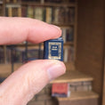 Load image into Gallery viewer, Hand holding a miniature book titled 'stock market' with a blurred bookshelf background