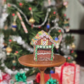 Load image into Gallery viewer, Decorative gingerbread house on a small wooden stand with a Christmas tree and presents in the background.