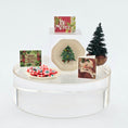 Load image into Gallery viewer, Christmas-themed display with cookies, cards, and a small tree on a white stand.