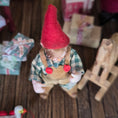 Load image into Gallery viewer, Clay elf character with a red hat and plaid shirt on a wooden surface with blurred gift boxes in the background