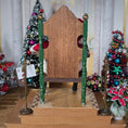 Load image into Gallery viewer, back of chair with green legs in a festive indoor setting with Christmas trees and decorations.