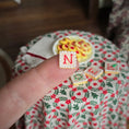 Load image into Gallery viewer, Hand holding a small square cookie with 'N' on it, with a decorative tablecloth in the background.