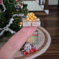 Load image into Gallery viewer, Miniature gold gift box with a bow held by a finger in front of a Christmas tree.