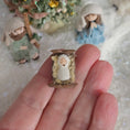 Load image into Gallery viewer, Miniature Bby Jesus in a manger held in a hand with other nativity figures in the background. miniature dollhouse