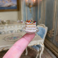 Load image into Gallery viewer, Miniature pastry held on a finger with ornate furniture in the background