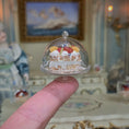 Load image into Gallery viewer, Miniature cake under a glass dome held by a finger with a decorative background