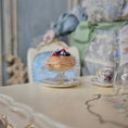Load image into Gallery viewer, Pastry on a small stand with a teacup and saucer on a table, blurred background