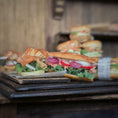 Load image into Gallery viewer, Sandwiches with salmon, lettuce, and onions on a wooden surface.