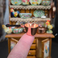 Load image into Gallery viewer, Small bronze pot held by a finger with a blurred background of ceramic dishes.