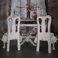 Load image into Gallery viewer, Two distressed white chairs with lace covers in front of a small table in a blurred indoor setting.