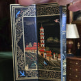 Load image into Gallery viewer, Decorative book with intricate illustrations of a tower and nature, held open by a hand.