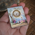Load image into Gallery viewer, Small decorative clock with a painted scene held in a hand on a wooden floor.