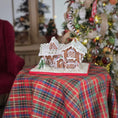 Load image into Gallery viewer, Gingerbread house on a plaid tablecloth with a Christmas tree in the background