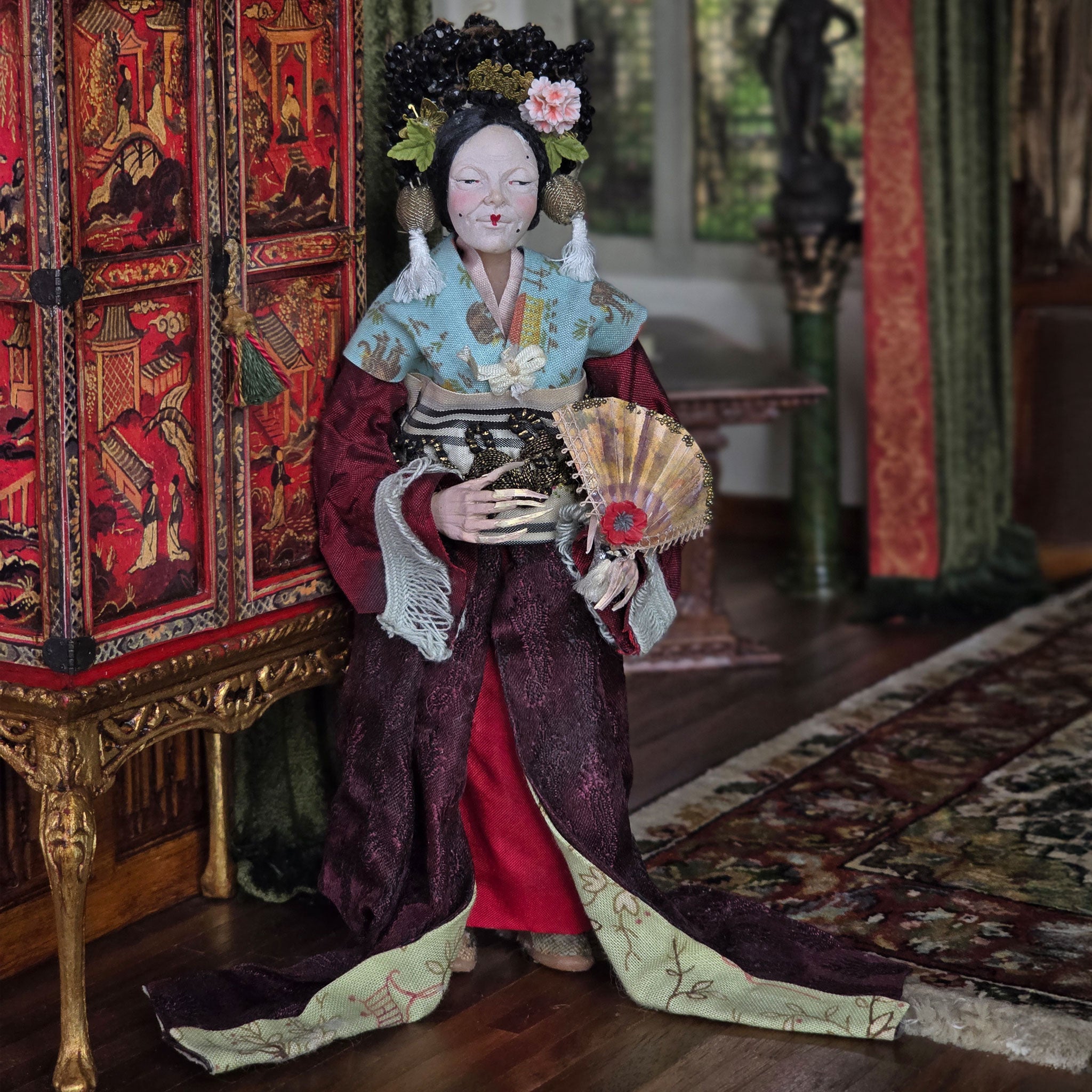 Traditional doll in ornate clothing holding a fan, standing in a decorated room.