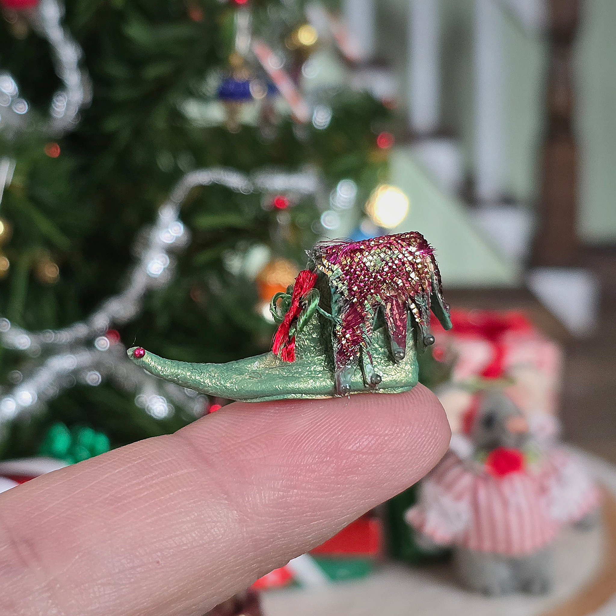 dollhouse miniature elf shoe held by a finger with a blurred Christmas tree in the background