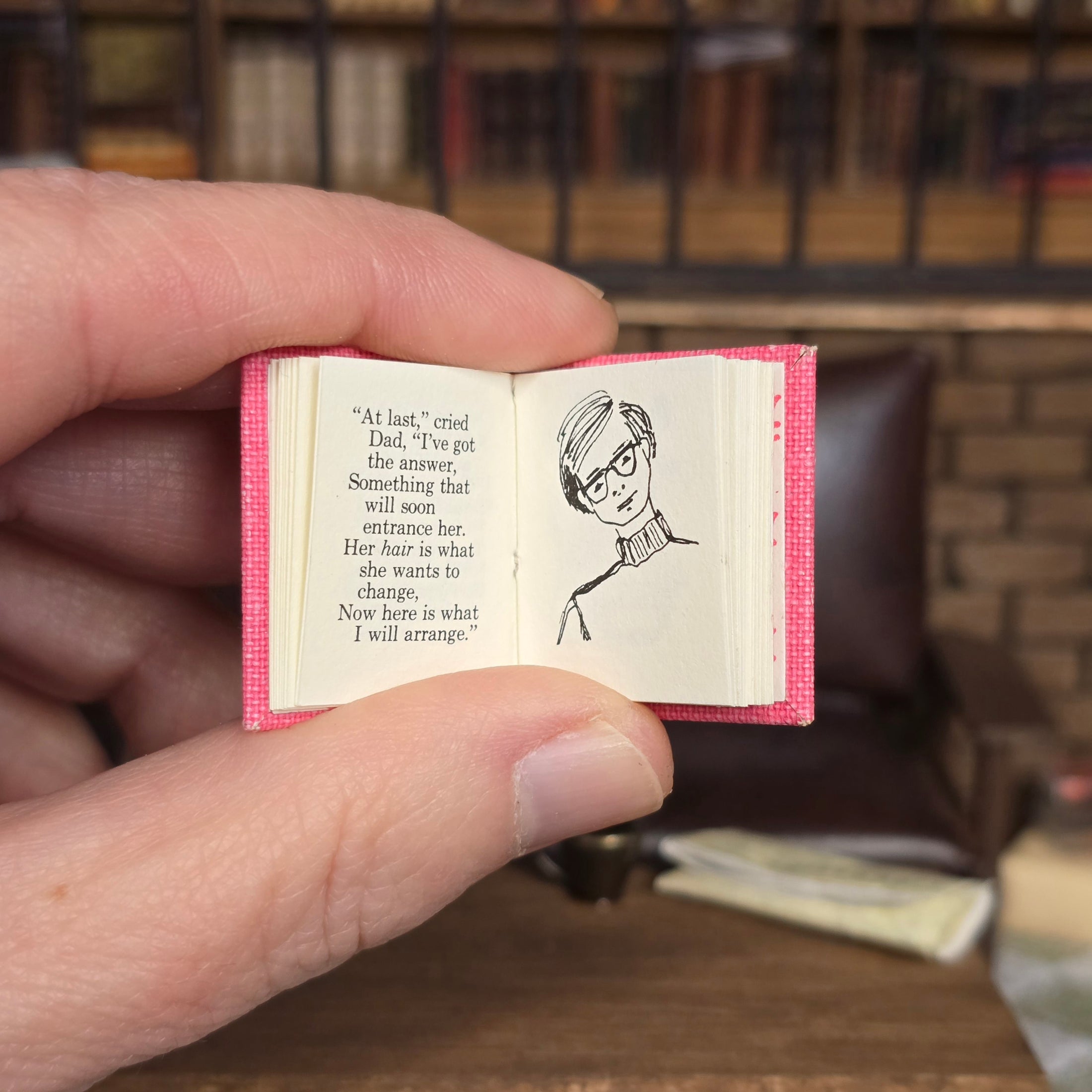 Hand holding a small open book with text and a drawing of a person wearing glasses, against a blurred bookshelf background.