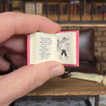 Load image into Gallery viewer, Hand holding a small open book with illustrations and text, set against a blurred indoor background.
