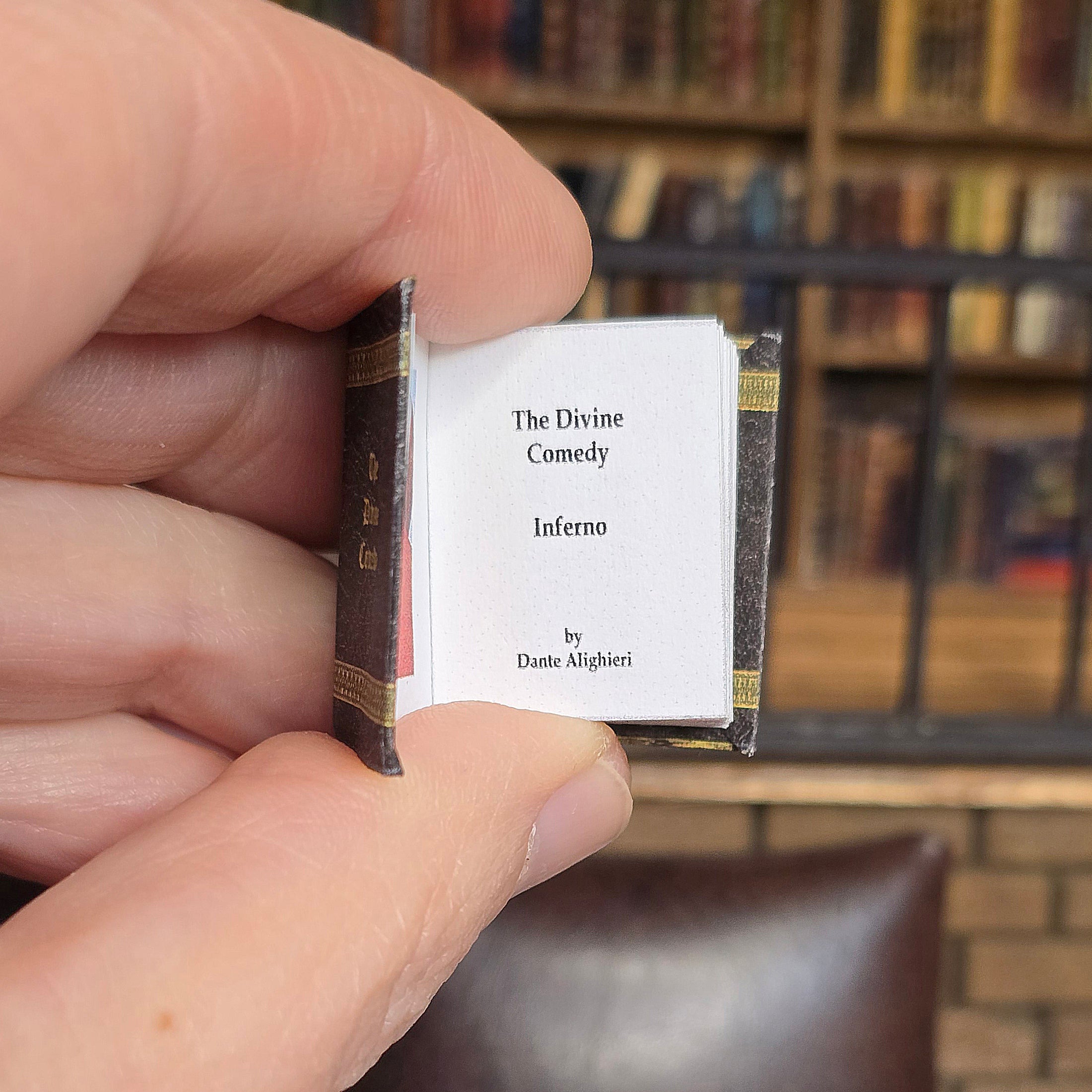 Hand holding a miniature book titled 'The Divine Comedy' with a blurred bookshelf in the background.