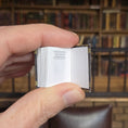 Load image into Gallery viewer, Hand holding a small open book with a blurred bookshelf background
