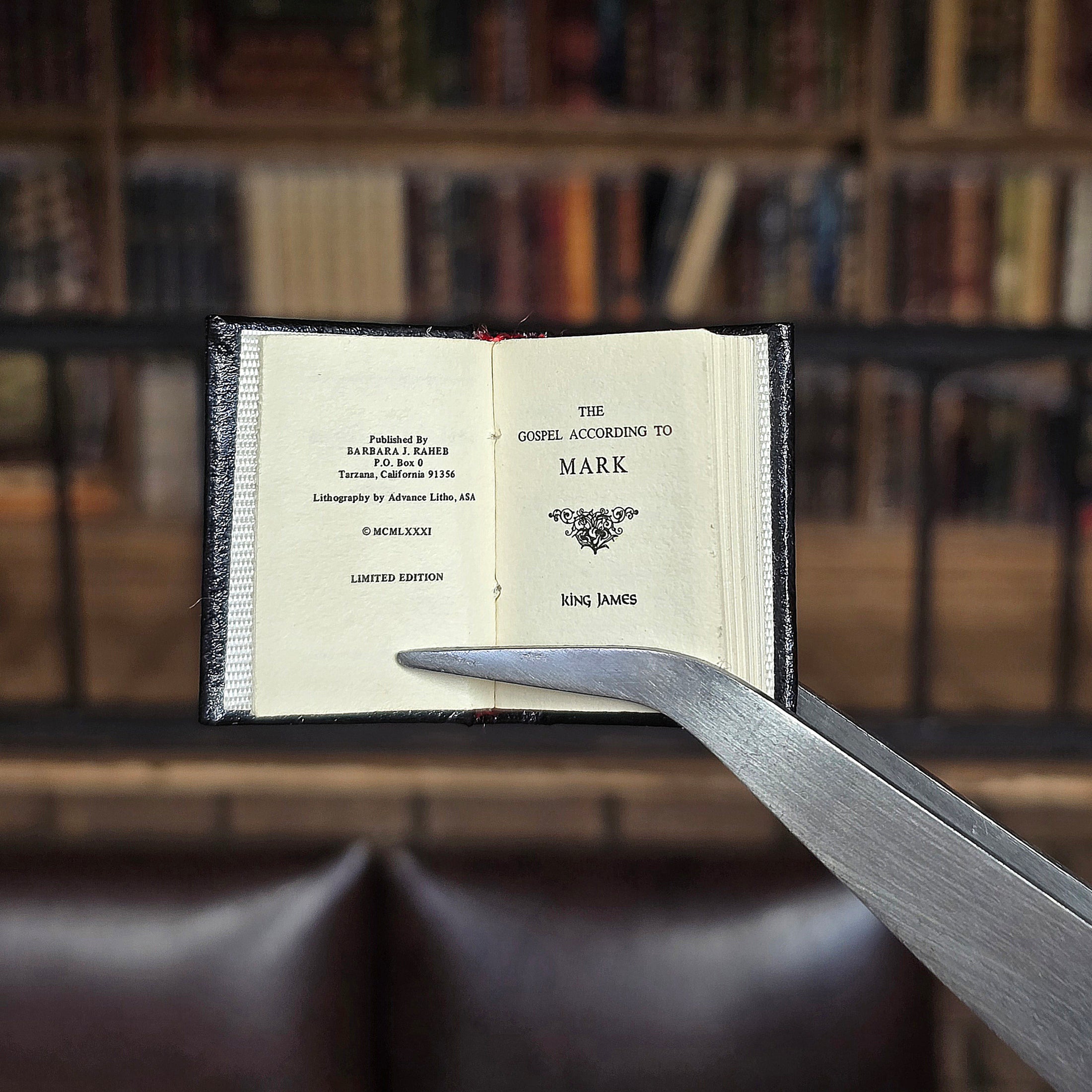 Miniature book titled 'The Gospel According to Mark' held open by tweezers with a blurred bookshelf background.