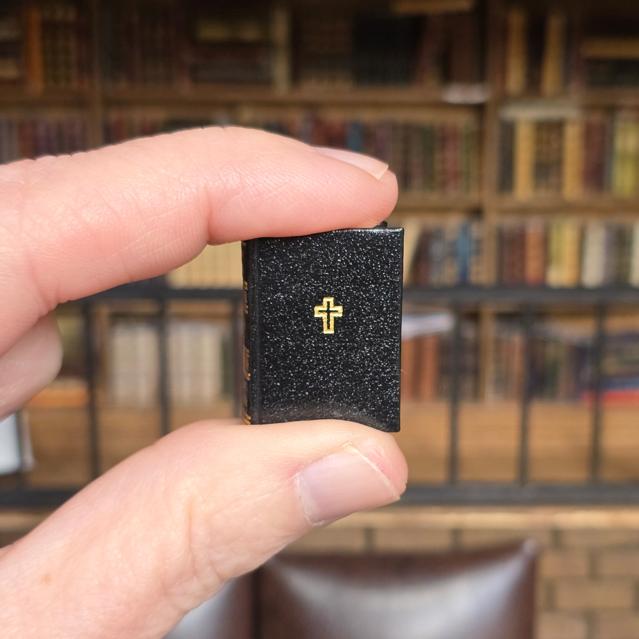 Hand holding a small black book with a gold cross in front of a blurred bookshelf.