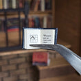 Load image into Gallery viewer, Small open book held by tweezers with text and a symbol, against a blurred bookshelf background.
