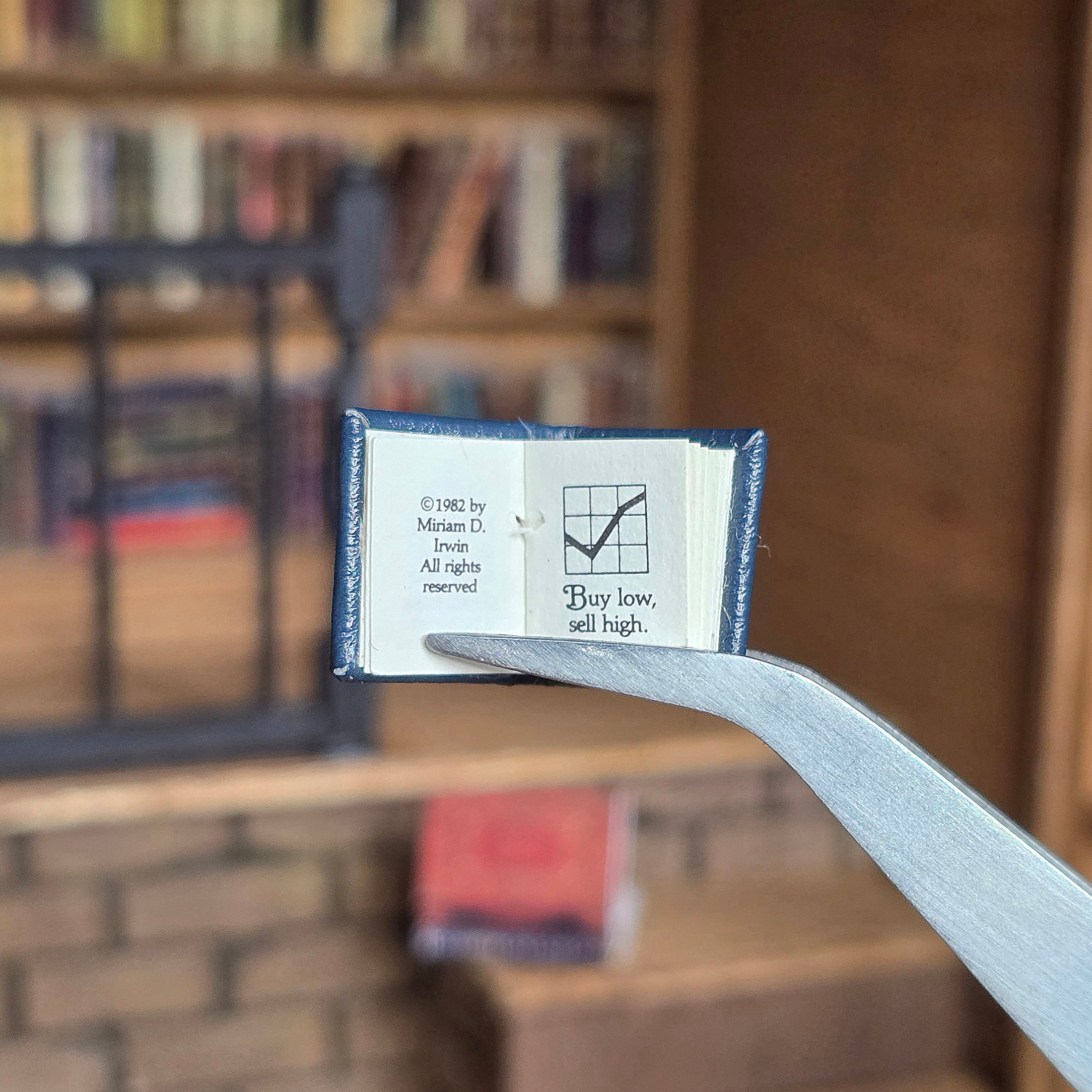 Small book held open by tweezers with a blurred bookshelf background