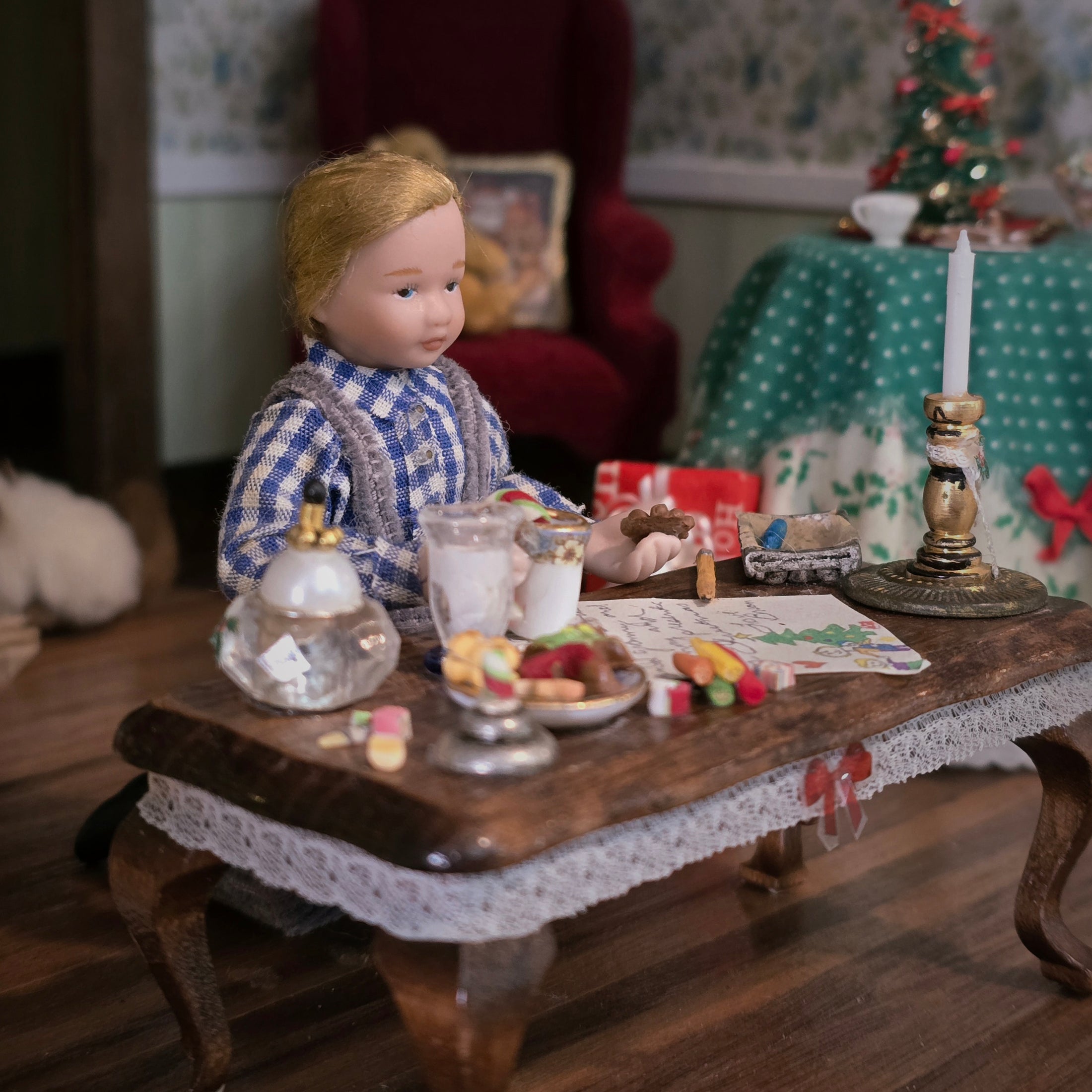 Doll in a checkered shirt sitting at a miniature table with various Christmas items in a cozy room setting.