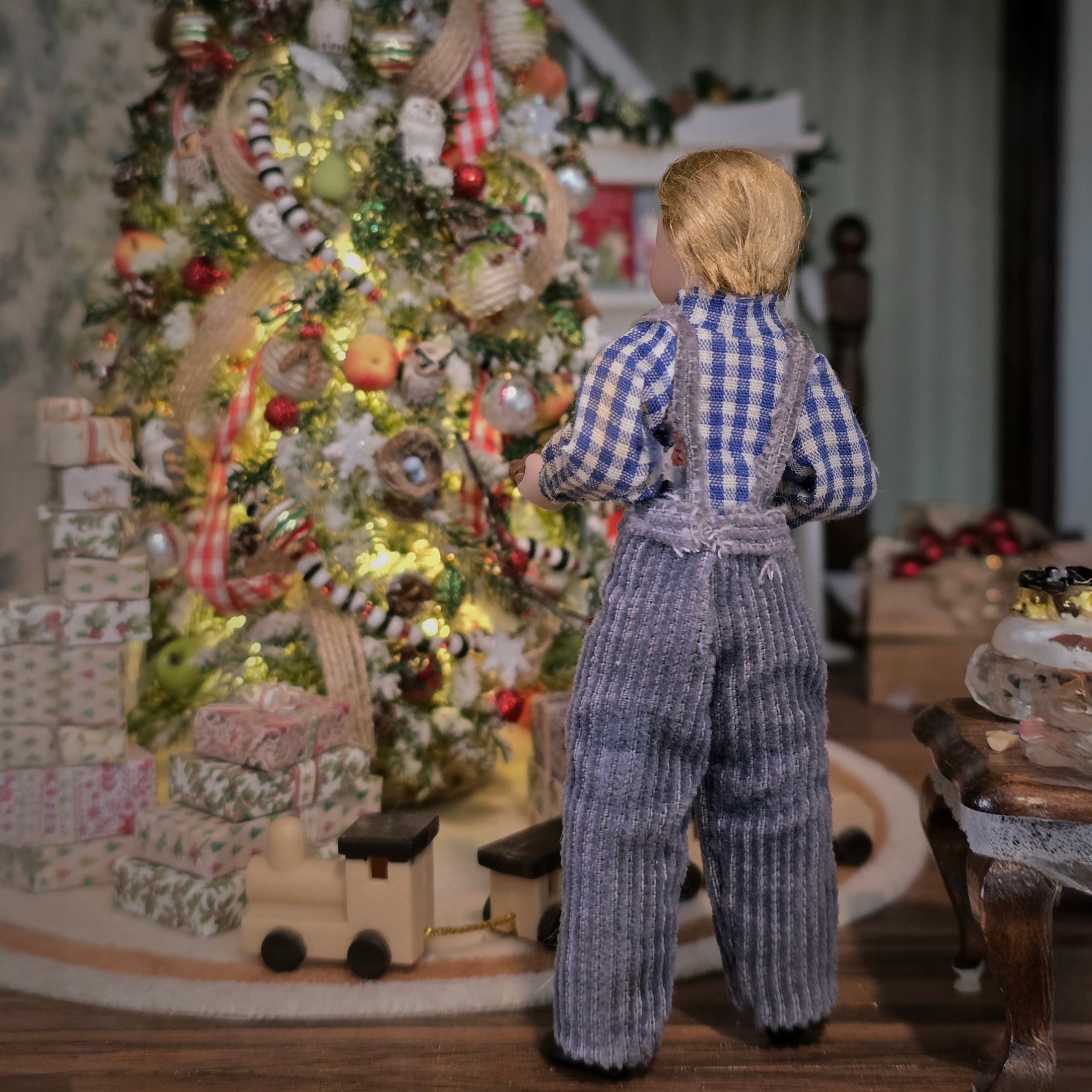 Doll in checkered shirt and overalls standing near a decorated Christmas tree with presents.