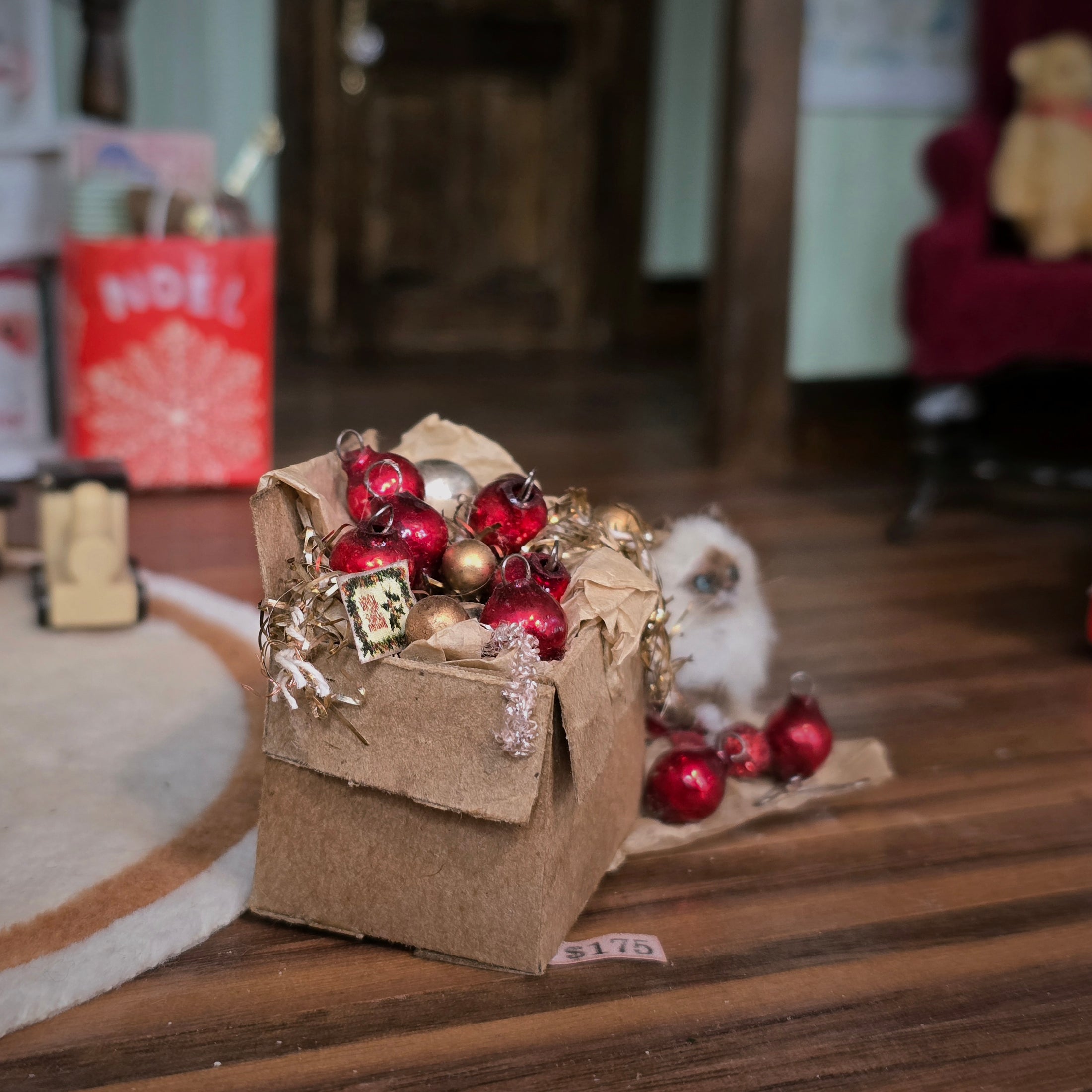 Small cat with Christmas ornaments in a cardboard box on a wooden floor.