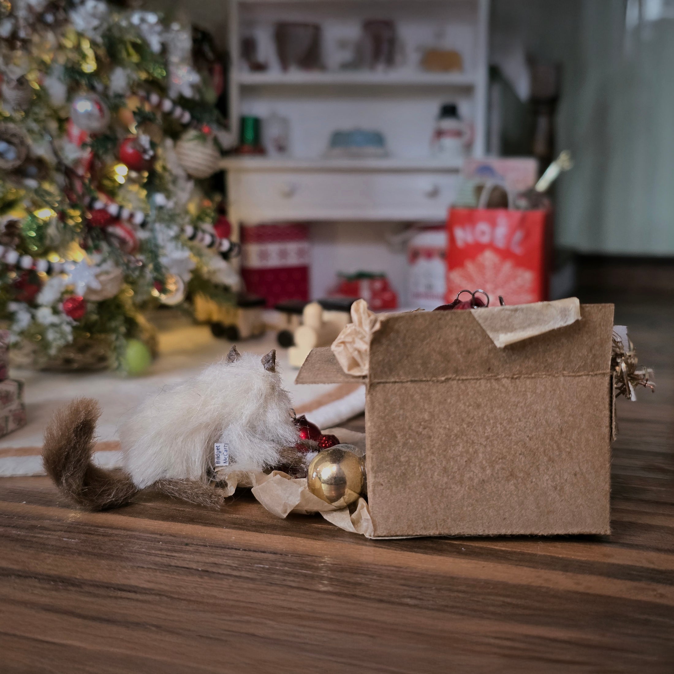 Small cat next to a cardboard box with Christmas decorations on a wooden floor. (back view)