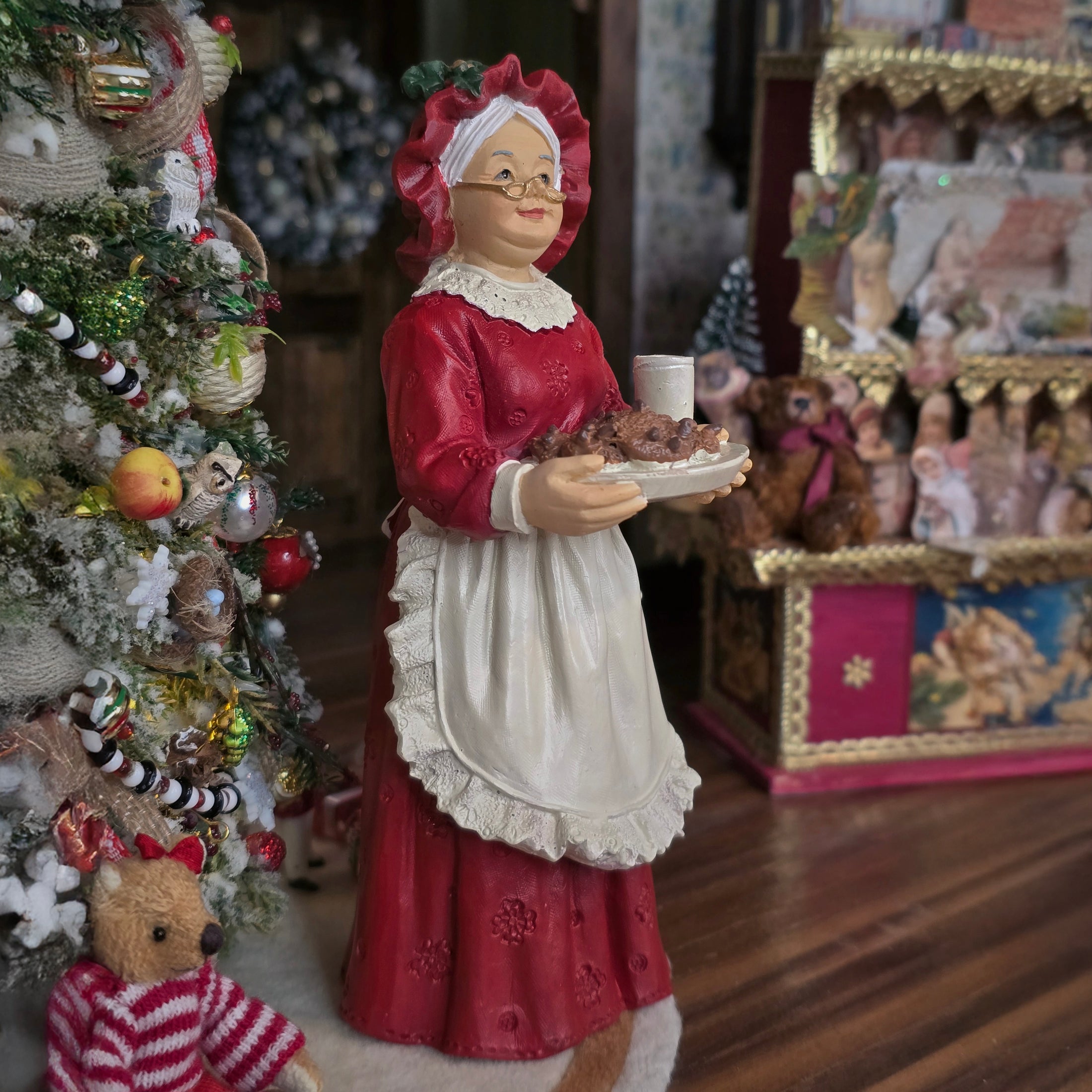 Mrs. Claus doll in a red dress holding a plate, with a Christmas tree and other decorations in the background.