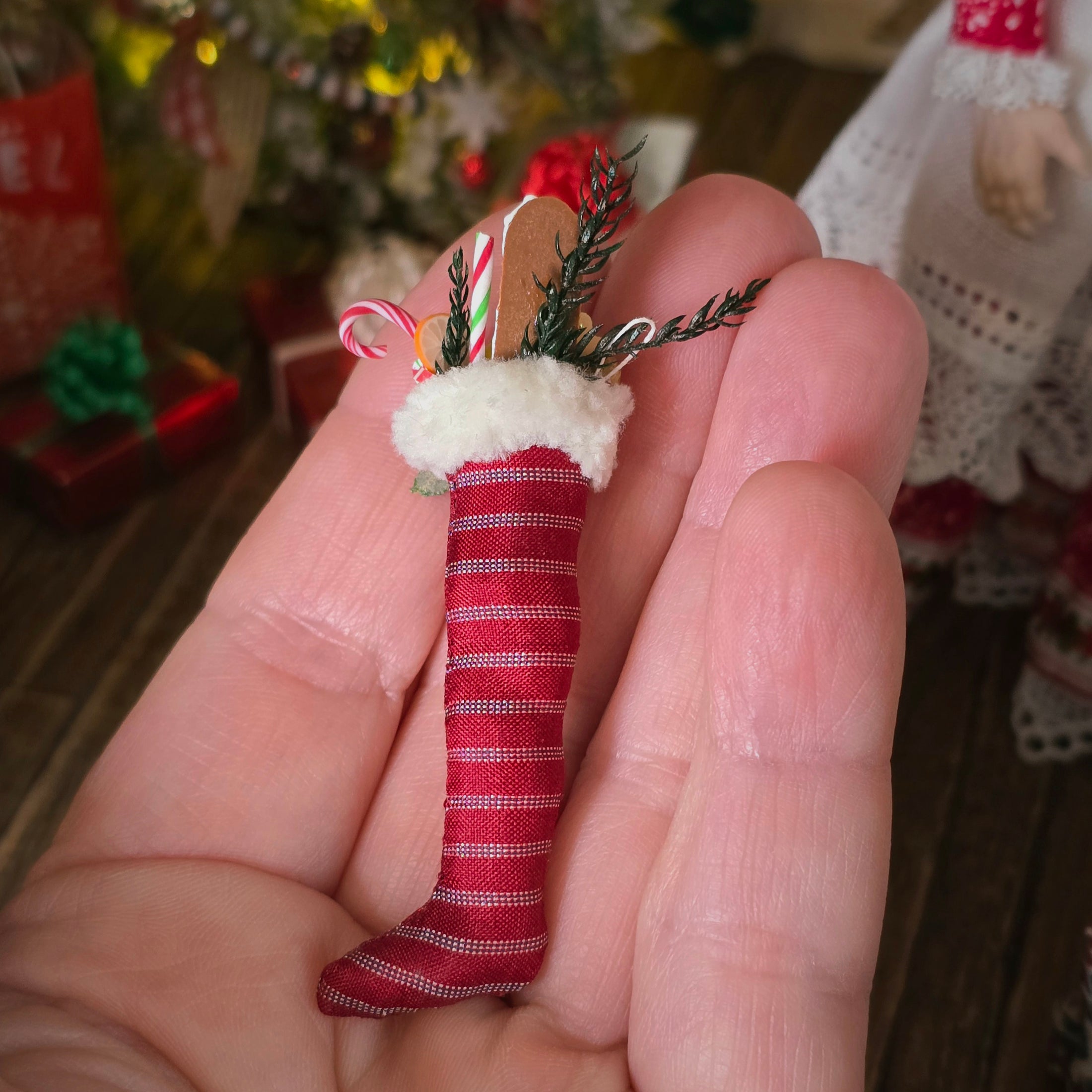 Small red and white striped stocking ornament held in a hand with a blurred Christmas tree and decorations in the background.