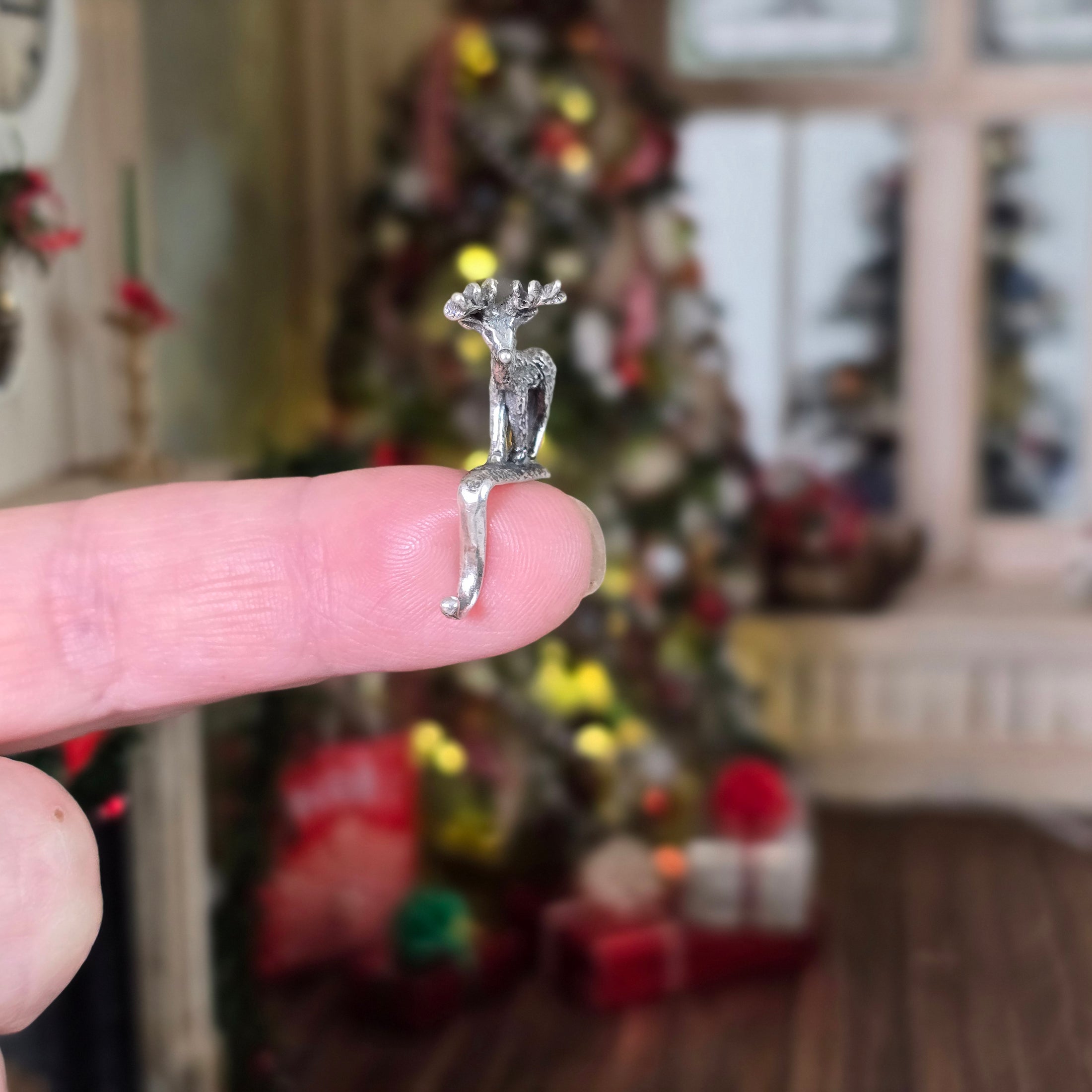 Miniature sterling reindeer stocking holder held on a finger with a blurred Christmas tree and decorations in the background
