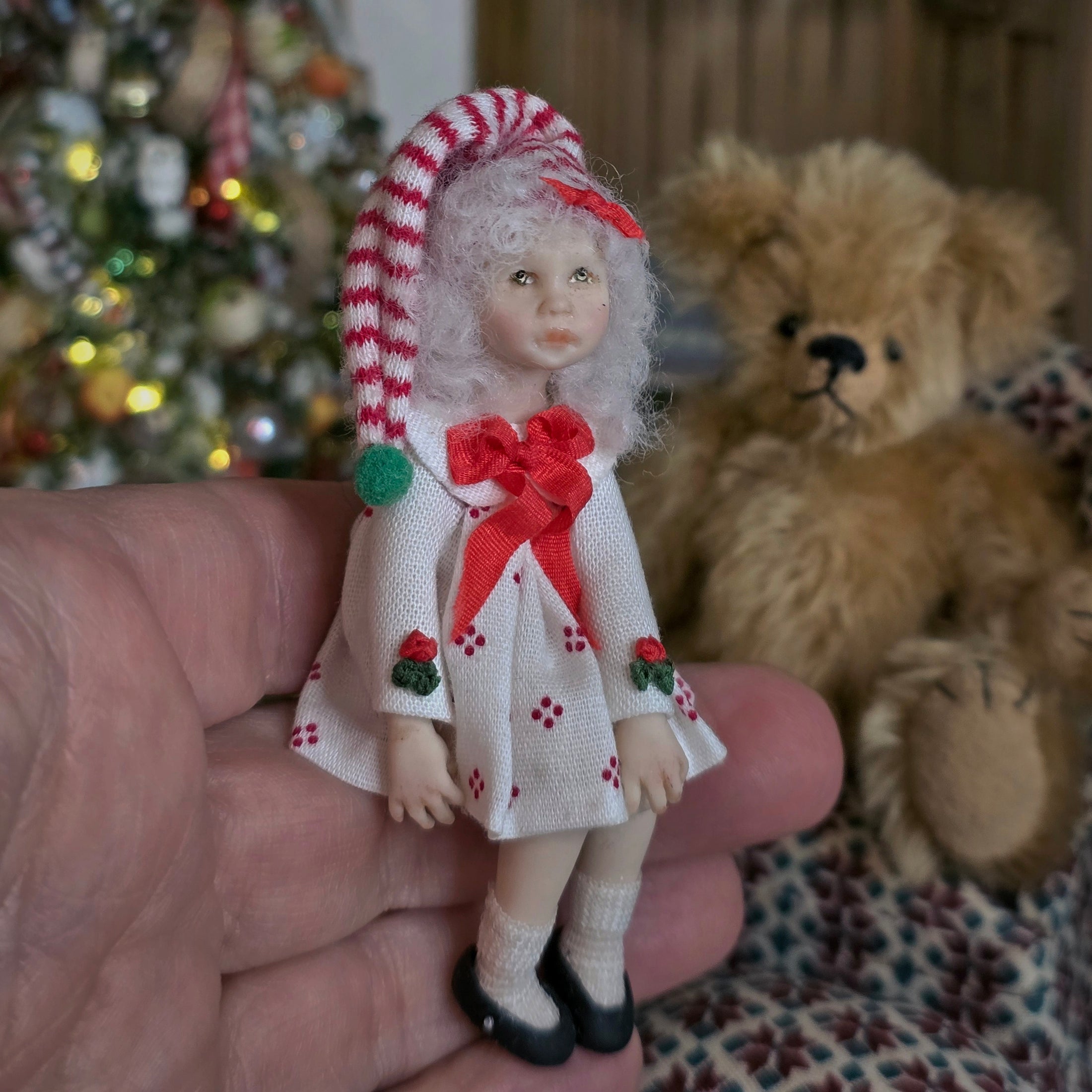 Small Christmas girl doll by artisan Carol McBride  with a candy cane hat and a teddy bear and Christmas tree in the background.