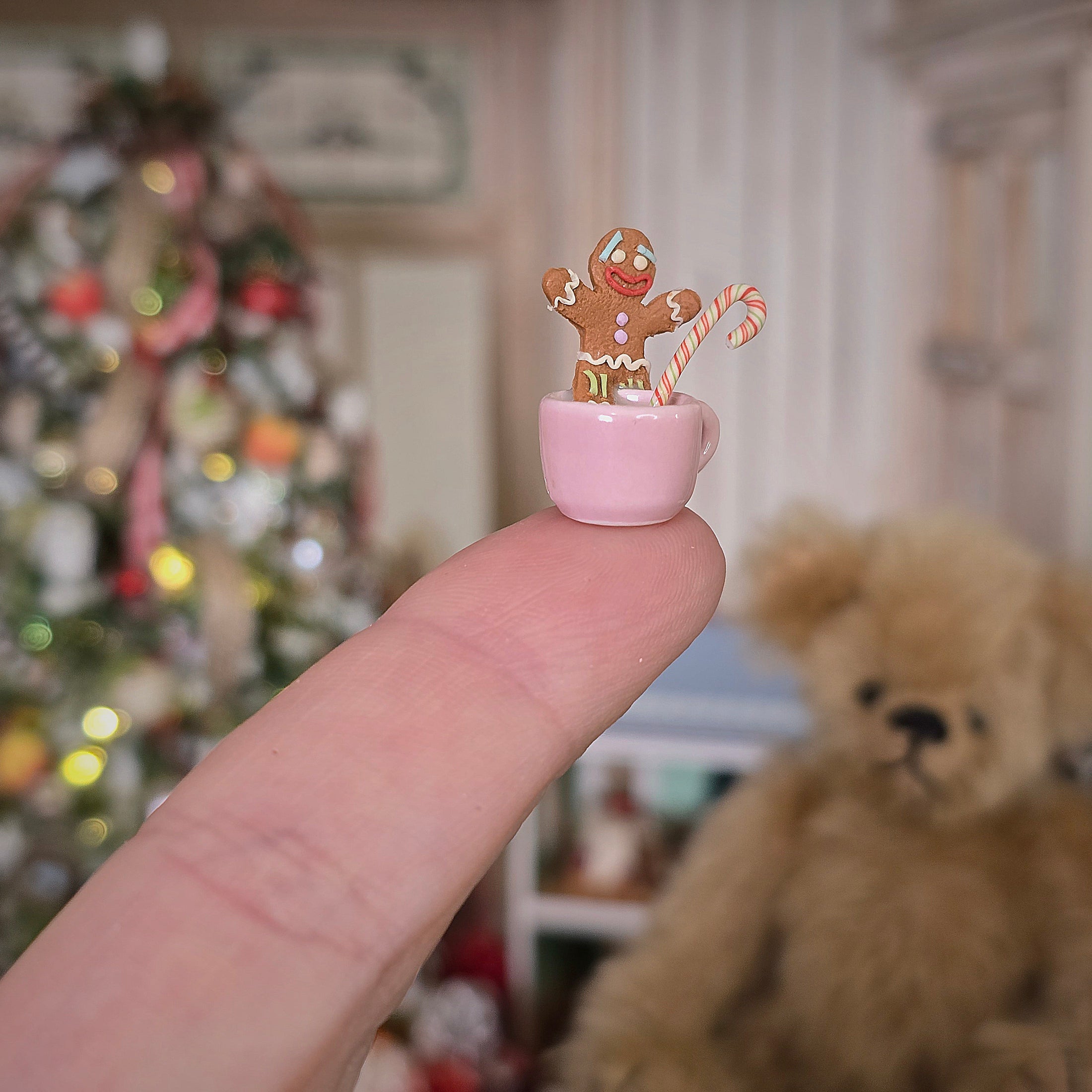 Miniature gingerbread man in a pink teacup held on a fingerswith a teddy bear and Christmas tree in the background.