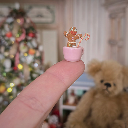 Miniature gingerbread man in a pink teacup held on a fingerswith a teddy bear and Christmas tree in the background.