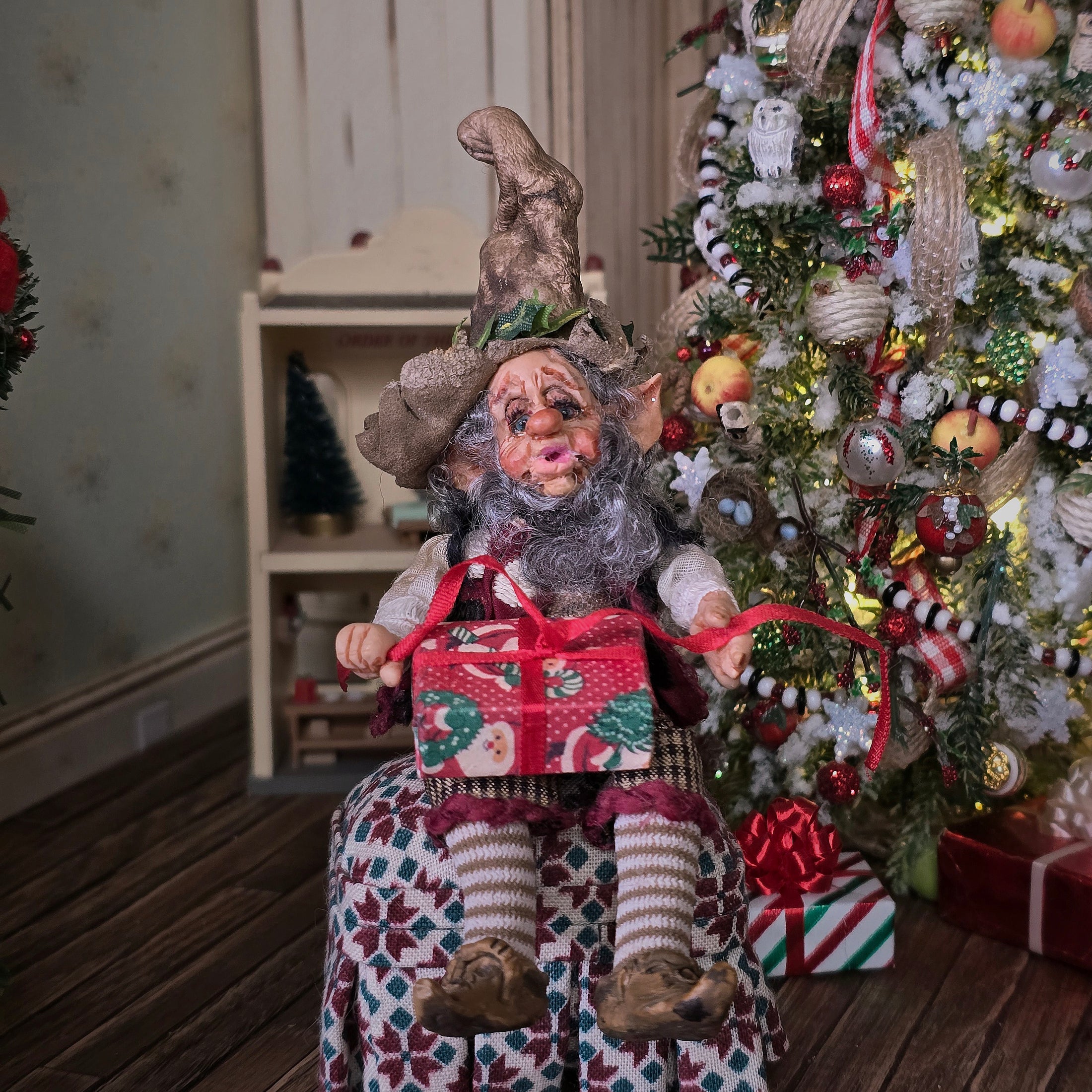 Decorative elf doll holding a gift in front of a Christmas tree.