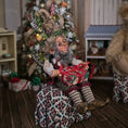 Load image into Gallery viewer, Decorative elf doll with a gift in a festive setting with a Christmas tree and presents.
