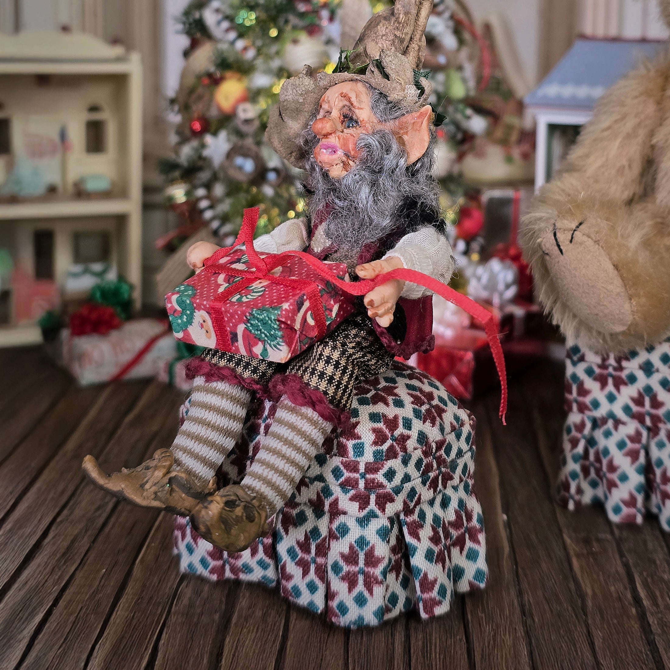 Decorative elf doll holding a gift in a festive setting with a Christmas tree and presents.