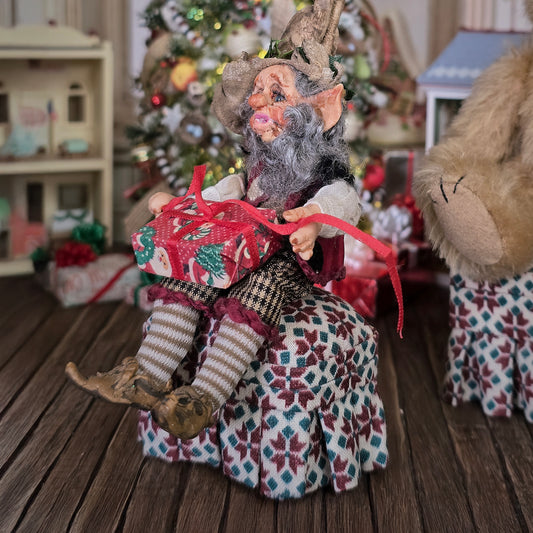 Decorative elf doll holding a gift in a festive setting with a Christmas tree and presents.