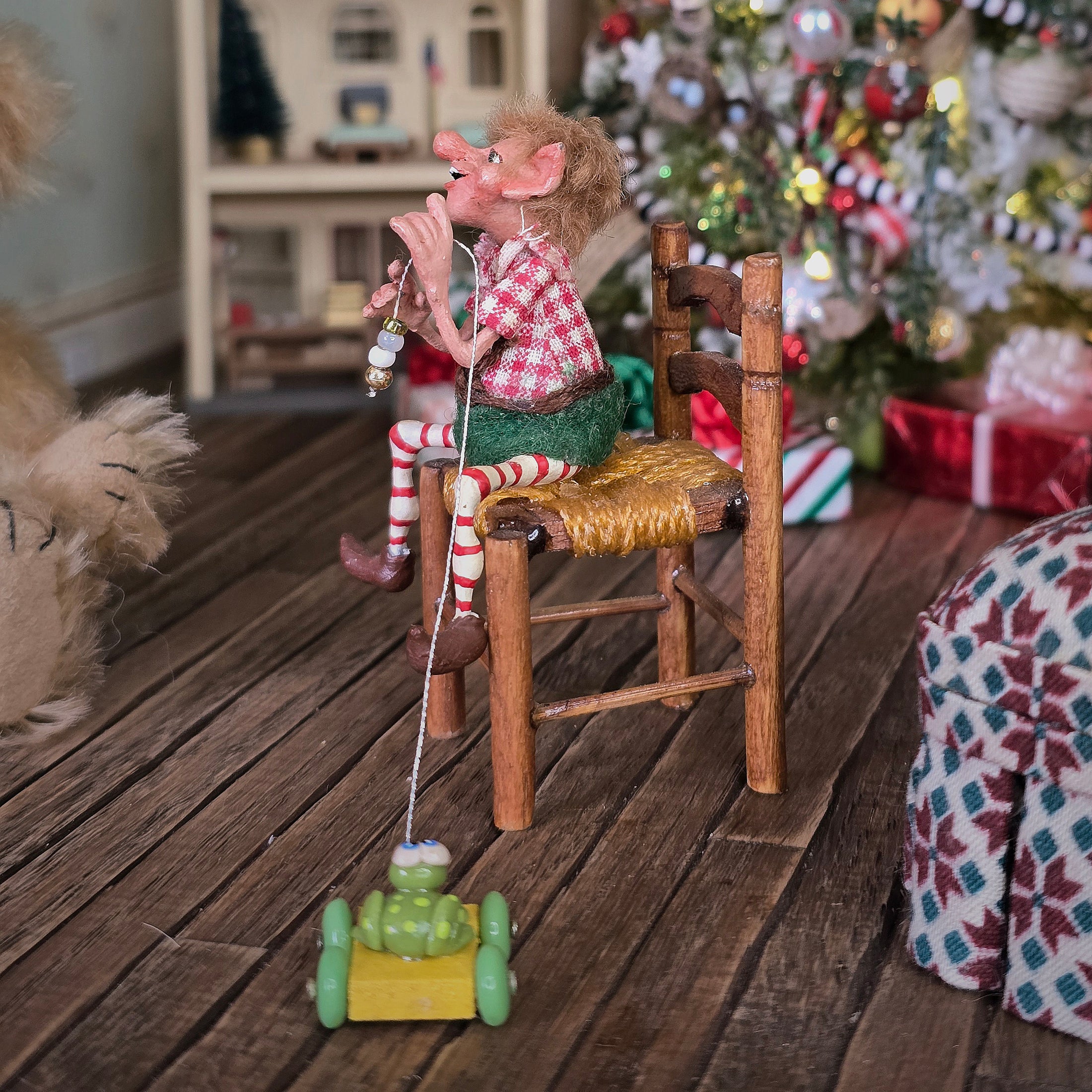 Grinning Elf Christmas Doll Cynthia Baron sitting on a chair with a toy car, in a festive room with a Christmas tree.