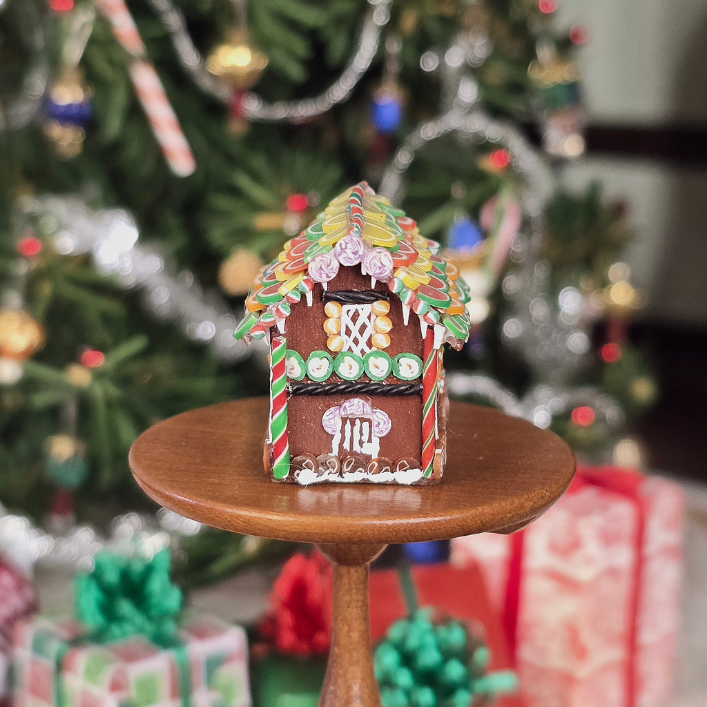 Decorative gingerbread house on a small wooden stand with a Christmas tree and presents in the background.