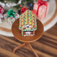 Load image into Gallery viewer, Colorful gingerbread house on a small wooden stand with Christmas decorations in the background.
