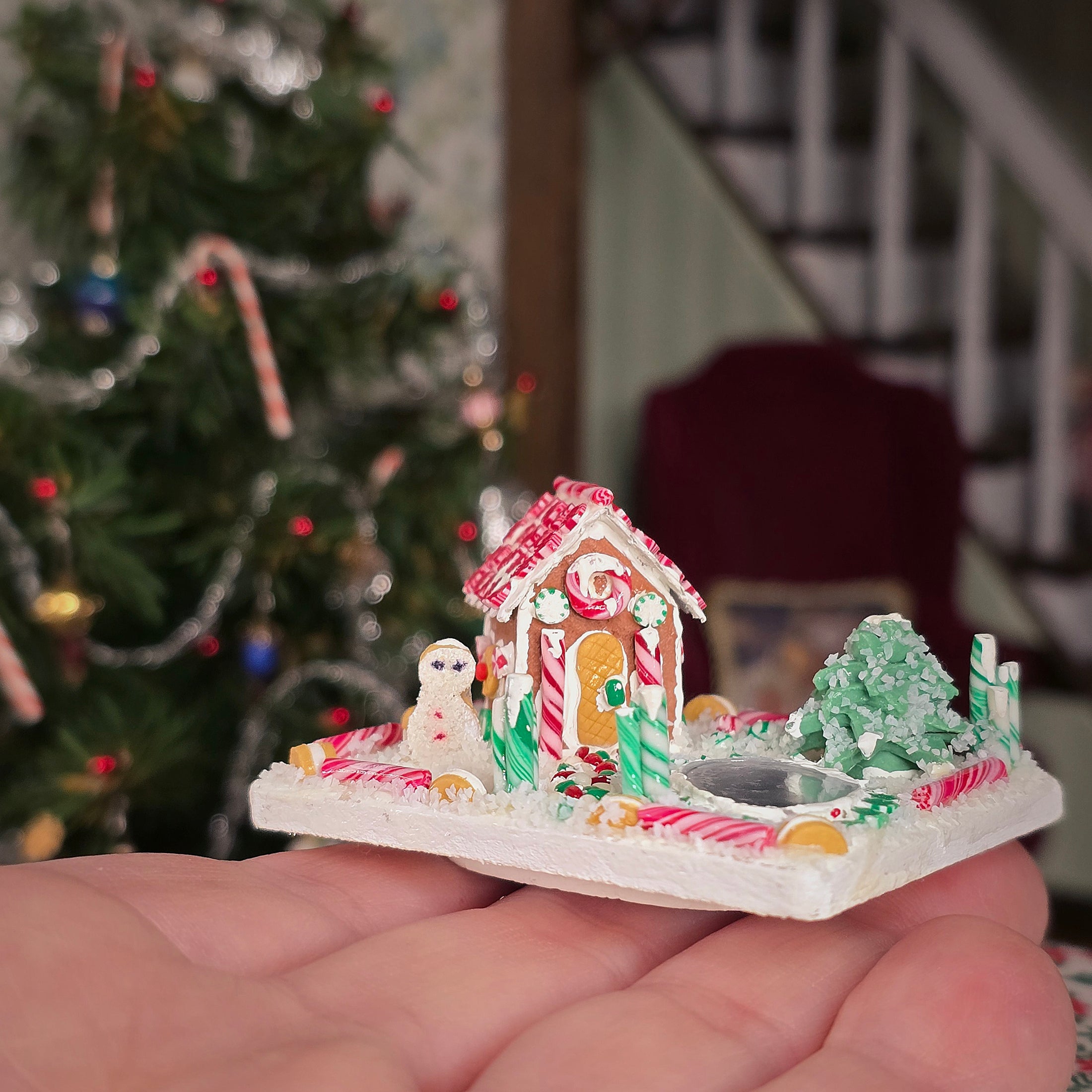 Miniature gingerbread house with candy decorations held  on a hand, in front of a Christmas tree.