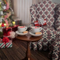 Load image into Gallery viewer, Two miniature cups with saucers on a small wooden table in a cozy room with a Christmas tree and patterned chair.
